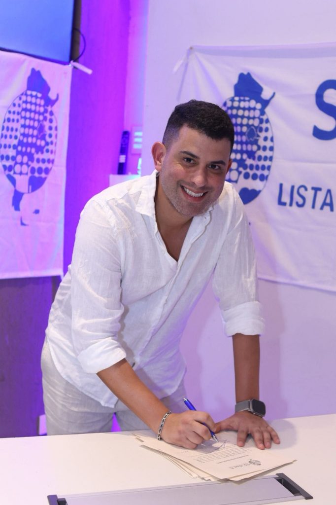 Man in a white shirt signing papers at a desk during an event, smiling at the camera with a purple-lit background and banner showing pineapples.